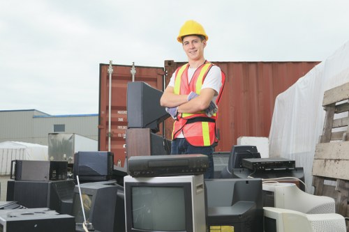 A young male worker standing outdoors on a gravel surface at a waste disposal yard, wearing a yellow safety helmet, high-visibility vest with orange and yellow reflective stripes, and casual clothing. He is smiling and has arms crossed, positioned behind a collection of discarded electronic appliances including computer monitors, printers, and other electronic waste stacked in front of him. In the background, there is a large open shipping container with a rust-colored exterior, partially filled with more waste materials. To the right, there are wooden pallets and stacked cardboard, suggesting a designated area for waste collection and sorting. The environment appears overcast, with diffuse natural light illuminating the scene, indicative of a typical day at a rubbish removal site serviced by companies like Commercial Waste Removal Coney Hall, operating within the local area near Coney Hall, possibly in the postcode area that includes surrounding towns.