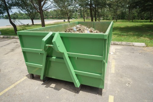 A large green metal skip bin filled with mixed rubbish including paper, plastic, and other debris, situated on an asphalt surface in an outdoor area with a grassy park in the background; the skip has reinforced sides with horizontal paneling and is positioned on a single section of a parking lot near trees, indicating a commercial waste collection site in or near Coney Hall. The environment is well-lit, and the bin appears ready for waste removal services provided by Commercial Waste Removal Coney Hall, emphasizing the handling of general rubbish for local businesses or residents.