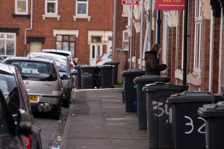 A row of traditional Victorian-style terraced houses with bay windows, pitched roofs, and brick chimneys, situated along a quiet residential street. In the foreground, two black waste or recycling bin